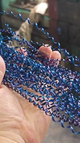 A hand holds a clump of curly blue metal shavings. The shavings are thin and curled into spirals. The hand appears to be an adult hand with visible skin texture. The background is out of focus and appears to be an industrial setting with some green and brown elements.