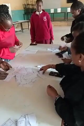 This video shows a group of individuals, likely students and possibly a teacher, gathered around a large table in what appears to be a classroom or common area. There are at least five people visible, with a young person in a red school uniform standing slightly apart. The majority of the individuals are focused on a pile of small, white papers scattered across the table. They are actively sorting, picking up, and examining these papers. Some individuals are using their mobile phones, possibly for reference or to capture information related to the papers. The environment appears to be a well-lit indoor space with a table, chairs in the background, and a whiteboard or bulletin board on the wall. The overall atmosphere suggests an activity involving sorting, organizing, or a group task related to the papers.
