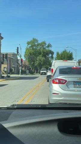 The video shows a view from inside a car, stopped at a red traffic light on a sunny day. A silver Chevrolet Cruze is directly in front. To the right is a Shell gas station and a Tobacco store. A man in a hat walks in front of the car toward the gas station. Other cars are stopped in the road ahead. Houses and trees line the street.