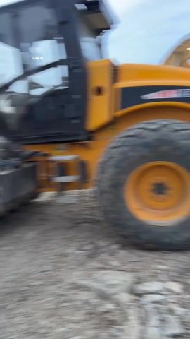 The video shows a construction site. Several pieces of heavy machinery are present, including an excavator, a steamroller, and another piece of equipment partially visible. A man wearing a high-visibility neon green jacket briefly enters the frame. The excavator is orange and black and is sitting on gravel. The video appears to be taken from a low angle, likely from someone standing or moving near the machinery. The sky is overcast.