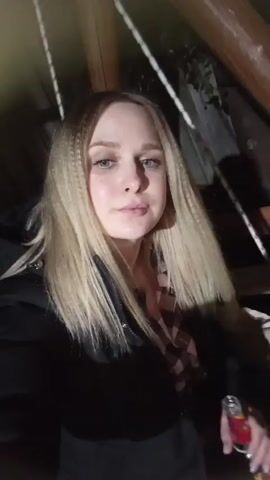 The video features a young woman with long, blonde hair that is partially crimped and partially straight. She has blue eyes and is looking directly at the camera with a slight smile. She is wearing a black jacket over a pink and white plaid shirt. In her right hand, she holds a small red and white bottle, from which she takes a sip at one point. The background is dark, suggesting it is night, but she is well-lit, possibly by a phone's flash or an artificial light source. Wooden structures and ropes are visible in the background, hinting at an indoor or covered outdoor setting, potentially a patio or gazebo. The woman appears relaxed and content. Russian speech and music are audible in the background. The location is Kauchuk Stantsiya, Russia.