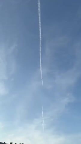 The media file is a vertical video showcasing a sky with several contrails from airplanes. The sky is a clear blue with some wispy white clouds scattered throughout, suggesting fair weather. The contrails appear as long, thin white lines against the blue background, indicating the flight paths of aircraft at high altitudes.

There are two prominent contrails that intersect, forming a cross-like shape in the upper portion of the frame. One contrail runs vertically down the center of the screen, and another runs horizontally across the top, meeting the vertical one. Further down the screen, another faint contrail is visible, running diagonally. The contrails are of varying thickness and clarity, with some appearing brighter and more defined than others, suggesting different aircraft or atmospheric conditions.

Towards the bottom of the frame, the edge of some greenery, possibly trees, is visible, hinting at a terrestrial location. The lighting suggests it is daytime, likely midday or afternoon, given the brightness of the sky. No people or specific objects are clearly discernible other than the contrails and natural elements. The overall mood conveyed is one of openness and the vastness of the sky. No visible text is present in the media file. The location context provided is Bönnigheim, Germany.