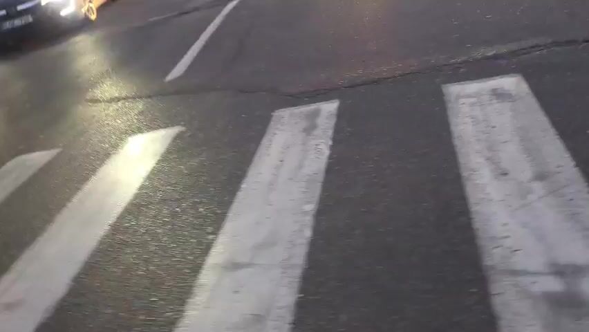 The video shows a street scene at dusk with a crosswalk in the foreground. Several cars are driving on the road, with their headlights on. One car is turning left, and another is driving straight ahead. There are also pedestrians visible, crossing the street. The sky is overcast, and the streetlights are on. The video appears to be shot from the perspective of someone walking or cycling across the crosswalk.