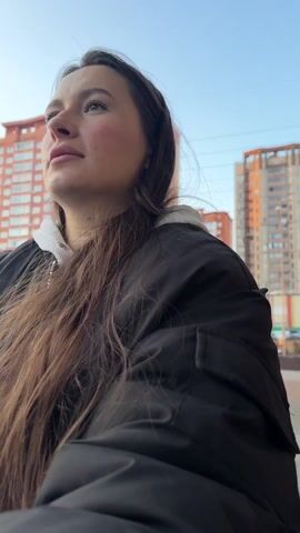 The video features a close-up shot of a young woman in an urban environment. She has long, straight, dark brown hair that falls over her shoulders. She is wearing a black puffer jacket over what appears to be a white hooded sweatshirt, with the white hood visible around her neck.

Throughout the video, she is looking towards her left (the viewer's right). Her facial expression is generally calm and contemplative, with a subtle, gentle smile appearing briefly around the 0:02 mark when she briefly closes her eyes. Her eyes are open for most of the clip, observing something off-camera.

The background consists of several multi-story buildings. Initially, prominent red/orange brick buildings with numerous windows are visible. As the camera subtly shifts or she moves, other buildings with modern, glass facades and some residential-style structures come into view. Parked cars, including a silver SUV, are also visible in what looks like a parking lot or street. The sky is bright, suggesting it is daytime, possibly late afternoon given the warm quality of the natural light. The overall setting is clearly an urban or suburban area. There are no other people prominently visible in the frame, though faint figures might be in the distance.