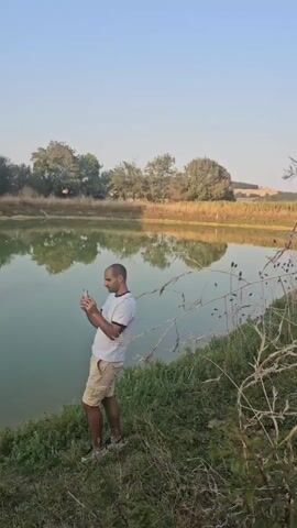 A man stands near the edge of a body of water, holding a smartphone. He is wearing a white t-shirt and beige shorts. Trees are visible in the background, and their reflections appear on the surface of the water. The sky is clear and blue. The man appears to be looking at or interacting with his phone.