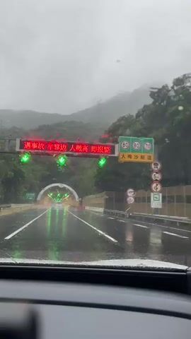 The video shows a view from inside a vehicle driving on a wet road towards a tunnel entrance. The sky is overcast and it appears to be raining, as evidenced by water droplets on the windshield and reflections on the road surface. A large electronic sign above the road displays red Chinese characters and green downward-pointing arrows, indicating a traffic advisory or instruction. To the right of the road, there are various traffic signs, including speed limit signs and warning signs. The tunnel entrance is brightly lit, with green lights illuminating the road leading into it. The surrounding environment is mountainous and covered in lush green trees. The road is divided into lanes, and there are barriers along the side. The video captures the car approaching and entering the tunnel.