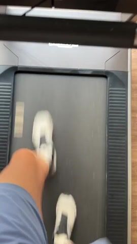 A person's legs and feet are visible, wearing blue shorts and white athletic shoes, as they run on a treadmill. The camera is positioned above and behind the runner, looking down at the treadmill belt and the runner's legs. The treadmill has a black frame and a gray belt. Above the belt, a red "STOP" button is visible. The floor around the treadmill appears to be light brown wood. The runner's legs are blurred due to the motion of running.
