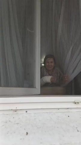 A woman with a cast on her left arm leans out of a window, speaking to someone off-camera. She is wearing a dark, shimmering shirt. She has dark hair and is smiling as she speaks. A sheer white curtain hangs behind her. The window is open. The scene appears to be outdoors, with natural light.