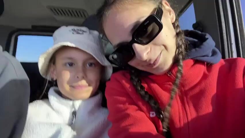 A short video clip shows three young women inside a vehicle. The woman on the right, in a red hoodie and sunglasses, is smiling and looking at the camera. Next to her, a younger girl in a white bucket hat is also smiling at the camera. To their left, another woman in a gray hoodie and a black hat is smiling at the camera. The interior of the vehicle appears to be a car or SUV. The background outside the vehicle is blurred but suggests an outdoor environment with some vegetation and possibly hills. The overall mood of the clip is cheerful and casual, suggesting friends or family on a trip.