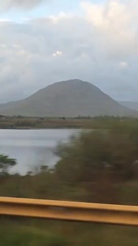 The video captures a scenic view from a moving vehicle, likely a car. The foreground is dominated by a blurred yellow guardrail and green foliage, indicating motion. Beyond this, a tranquil body of water, possibly a lake or a wide river, stretches across the middle ground. In the background, a large, rounded mountain rises against a cloudy sky, which transitions to a clearer blue near the horizon. The landscape appears to be rural or mountainous, with sparse vegetation visible on the lower slopes of the mountain and around the water's edge. The overall impression is one of a journey through a natural, serene environment.