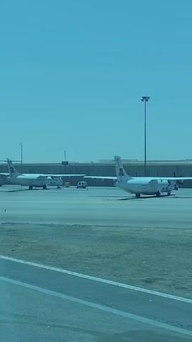 The video captures an outdoor scene at an airport apron under a clear, bright blue sky, suggesting daytime. Two white Air Cairo turboprop aircraft are prominently visible, parked on the asphalt.

The aircraft on the right, bearing the registration "SU-BLM", is positioned with its tail facing slightly towards the viewer, clearly displaying the "AIR CAIRO" livery on its fuselage and tail fin. Its propellers are stationary.

Further in the background, to the left, another Air Cairo aircraft, registered "SU-BLN", is also parked. A small white ground support vehicle is situated between the two aircraft.

The foreground shows a concrete or asphalt surface with painted lines. Tall light poles are scattered across the airfield, and a low building or wall is visible in the distant background. The overall impression is a sunny day at an airport, likely in Cairo, Egypt, given the airline and context.
