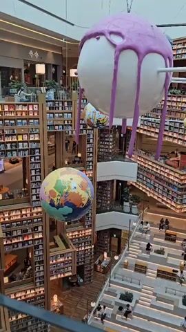 The video provides a high-angle view of a multi-level library with an atrium. Large, spherical decorations are suspended from the ceiling, one of which resembles a white orb with purple icing dripping down its sides. Another sphere appears to be a globe. Bookshelves line the walls of each level, filled with numerous books. A wide, tiered staircase with seating areas is visible in the center of the atrium, with many people seated on the steps. The lighting suggests it is daytime, with natural light entering from above. The overall atmosphere is one of a spacious and modern library.