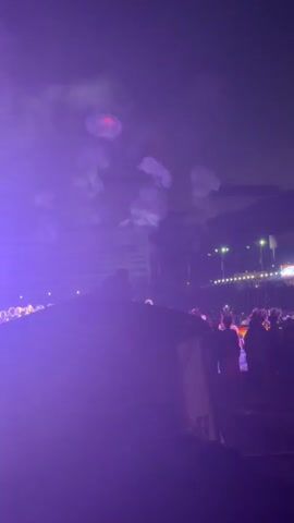 The video shows a crowded outdoor event at night. Purple lights illuminate the sky, creating a smoky atmosphere. A stage or raised platform is visible in the background, with several figures on it and what appear to be fire effects. A crowd of people is gathered in front of the stage. The perspective suggests the video is being filmed from within the crowd.