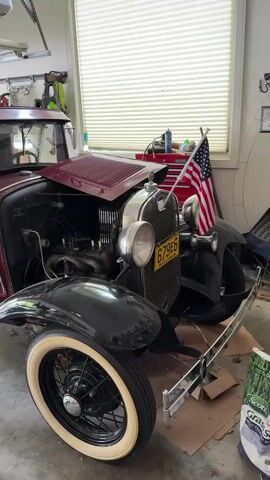 The video shows a maroon Model A Ford car parked in a garage. The car has black fenders, a black top, and whitewall tires. The hood is open, revealing the engine. An American flag is sticking out of the front of the car, and a license plate is visible. Various tools and equipment are visible in the garage, including a garage door opener, a red toolbox, and bicycles. The garage door is partially open, revealing the outdoors.