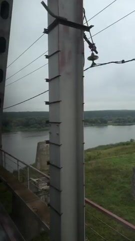 The video pans across a view from a bridge. The bridge is made of large metal beams with some visible rust. There are power lines overhead. In the distance, a wide river is visible with trees on the far bank. The sky is overcast. There is a concrete structure on the riverbank, possibly a dam or pier. The camera moves to show a tall, cylindrical concrete tower with a platform at the top, also on the riverbank. A metal staircase leads up to this platform. The view continues to show the river and the opposite bank.