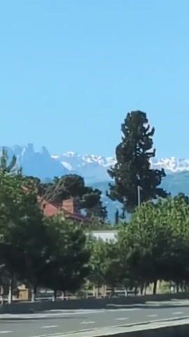 The video shows a roadside scene with mountains in the background. In the foreground, there is a road with cars driving on it. There are trees and streetlights along the side of the road. The sky is clear and blue.