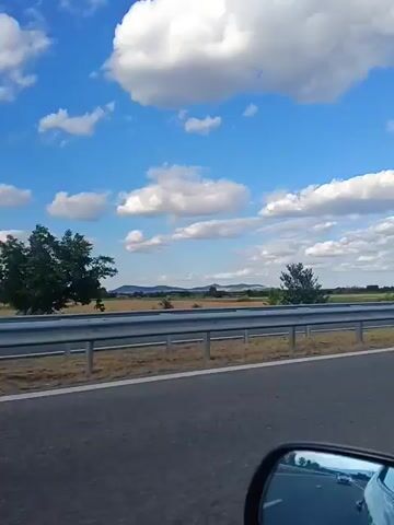 The video is filmed from a moving vehicle, looking out the passenger window. The scene is a highway with a metal guardrail separating the road from a dry, grassy field. In the distance, there are trees and rolling hills under a bright blue sky filled with fluffy white clouds. A billboard featuring two people, one of whom is wearing sunglasses, is visible on the right side of the frame. The reflection in the car's side mirror shows other vehicles on the highway. The overall impression is a sunny day travel scene.