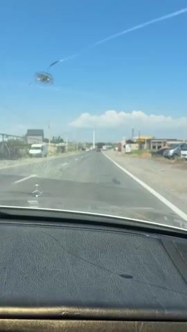 The video captures a view from inside a car, looking out through the windshield. The scene is a road in Yeraskh, Armenia, on a bright, sunny day. The sky is a clear blue with a few scattered white clouds. The road appears to be a two-lane highway with a white dividing line.

On the left side of the road, there are some buildings, including what looks like a house and a smaller structure, as well as a white van parked. Further down the road on the left, there are more buildings and a tall tower. On the right side of the road, there are more commercial buildings, including a gas station with a sign that reads "PETROL" and another sign with yellow and red text. There are also some cars on the road, and the car the video is being filmed from appears to be in motion.

There are some smudges and what appears to be a bug or debris on the windshield, obscuring some parts of the view. The dashboard of the car is visible in the foreground, made of black textured material. The overall impression is one of a typical day of driving through a semi-urban or roadside area in Armenia. The time of day is clearly daytime, given the bright sunlight and clear sky, suggesting it could be late morning or early afternoon. There are no people visible within the frame.