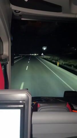 The video shows a view from inside a vehicle, possibly a bus or a truck, driving at night. The camera is positioned behind the driver's seat, facing forward through the windshield. The road ahead is illuminated by the vehicle's headlights, revealing two lanes separated by white dashed lines. The surrounding environment is dark, with only faint lights visible in the distance and along the side of the road. A triangular warning sign with a yellow background is visible on the right side of the road. The interior of the vehicle shows a dashboard with some instruments and a screen, as well as parts of seats. The footage is shaky, suggesting movement.