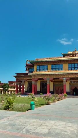 The video pans across a large, ornate building with Tibetan architectural features. The building is yellow with red and gold accents. It has multiple windows on the upper floor and large red doors at the entrance. In front of the building is a paved courtyard with a grassy area on the right. Potted plants with pink flowers line the walkway. Two trash cans, one green and one blue, are visible in the courtyard. The sky is a clear blue with a few white clouds.