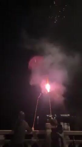 Two people are standing on a balustrade at night, setting off fireworks. The fireworks shoot up into the air, leaving trails of smoke. The night is dark and clear, with some lights visible in the distance. The people are silhouetted against the night sky. One is wearing a hooded jacket and the other is wearing a dark jacket. They are both looking up at the fireworks. The balustrade is ornate and made of concrete.