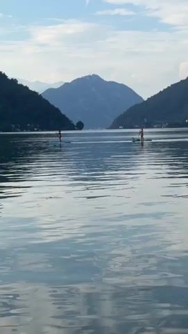 The video shows a wide shot of a tranquil lake surrounded by lush green mountains under a hazy, partly cloudy sky. The water is calm with gentle ripples reflecting the sky and surrounding landscape. Two individuals are visible in the distance, each on a stand-up paddleboard. The person on the left appears to be a woman, and the person on the right also appears to be a woman. They are both positioned in the middle of the lake, with the mountains forming a dramatic backdrop. The overall atmosphere is peaceful and serene, suggesting a leisurely outdoor activity.
