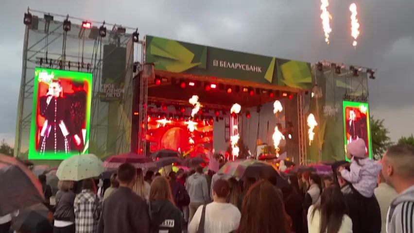 A live music concert is taking place on an outdoor stage. A crowd of people is gathered in front of the stage, many holding umbrellas due to rain. The stage is elaborate with a large backdrop featuring the logo "БЕЛАРУСБАНК" and green geometric designs. Two large LED screens flank the stage, displaying images of a woman singing. The stage itself is illuminated with numerous red lights, and a circular graphic resembling the sun or moon is visible on the central screen. Musicians are performing on stage, including a drummer on the left side of the stage. The sky appears cloudy, suggesting overcast weather.