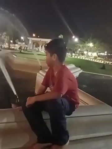 The video shows a young boy wearing a red shirt and dark pants sitting on the edge of a fountain at night. Water is spraying from multiple fountains in the background, creating a misty effect. The boy is looking away from the camera, towards the fountains. The environment appears to be a park or a public space with lights and buildings visible in the distance.
