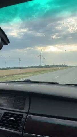 The video shows a view from inside a car as it drives on a highway. The dashboard of the car is visible in the foreground, with a screen displaying information. The windshield reveals a cloudy sky with sunlight breaking through in the distance. A field of crops is on the left side of the road, with a wind turbine visible further down. There are also utility poles lining the road. A white van drives past in the opposite direction. The video is shot from a passenger's perspective, looking forward through the windshield.