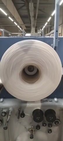 This is a close-up, static shot of a large spool of white thread or yarn on an industrial machine. The spool is spinning rapidly, causing the threads to appear blurred. The machine itself is industrial, with metal components visible. The background is out of focus but appears to be a factory setting with overhead lighting and pipes. There are no people visible in the video.