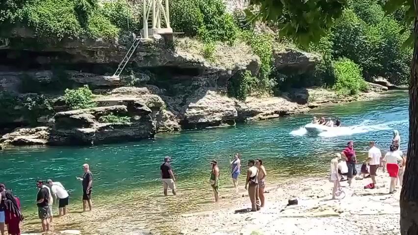 The video shows a group of people on a rocky, sandy shore next to a clear blue river. A small white speedboat with several people on it is moving at speed across the river, leaving a white wake. Some people are standing in the shallow water at the edge of the river, while others are standing or sitting on the shore. In the background, there are steep, rocky cliffs covered in lush green vegetation. There is also a white structure with a ladder leading up it visible on the cliff face. Sunlight is bright, indicating it is daytime and likely warm weather.
