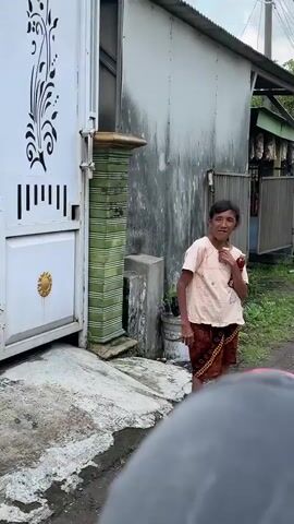 A person stands outdoors in front of a building and a white gate. The person is wearing a light-colored t-shirt and a patterned skirt. The person is looking at the camera and smiling. The building behind the person is white and appears to be made of corrugated metal. The gate is white and has a decorative pattern. The ground is paved with concrete.