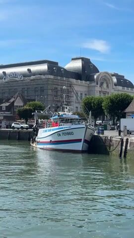 A fishing boat with the identification number CN 721860 is docked by a harbor wall. The boat is white with red and blue stripes along its side. In the background, a large building with a dark, mansard roof is visible, along with other smaller buildings and trees. A few people are walking along the waterfront, and a white van is parked near one of the buildings. The sky is blue with some scattered clouds, and the water in the harbor is green. A seagull is flying in the sky. The scene appears to be a sunny day in a coastal town.