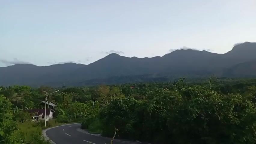 The video shows a winding road in a lush, green area with mountains in the background. The mountains are dark and silhouetted against a pale blue sky. There are some clouds surrounding the peaks. In the foreground, there's a mix of trees and foliage. A single-story house can be seen partially obscured by the trees.  A streetlight stands alongside the road.