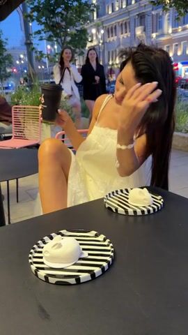 A young woman with long dark hair, wearing a white dress, sits at an outdoor cafe table. She holds a black to-go cup and drinks from it through a straw. Her left leg is extended, and her right arm rests on the table. In front of her are two small white desserts on black and white striped plates. In the background, two other women in white and black outfits walk past, and other people are visible at tables and in the street. The setting appears to be a city street at dusk, with buildings and streetlights illuminated.