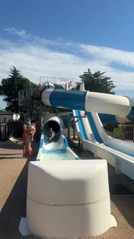 A child is sliding down a blue and white water slide at a water park on a sunny day. The child is about to splash into the pool at the bottom of the slide. There are people standing on the side of the slide watching. The slide is elevated and made of plastic. The sky is blue with some white clouds.