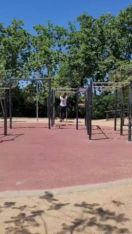 The video shows a man exercising on a pull-up bar. He is wearing a white t-shirt, black pants, and tennis shoes. He is hanging from the bar with both hands. There is a yellow resistance band attached to the bar. The man is in an outdoor exercise park with red flooring and other exercise equipment nearby. There are trees in the background and the sky is blue.