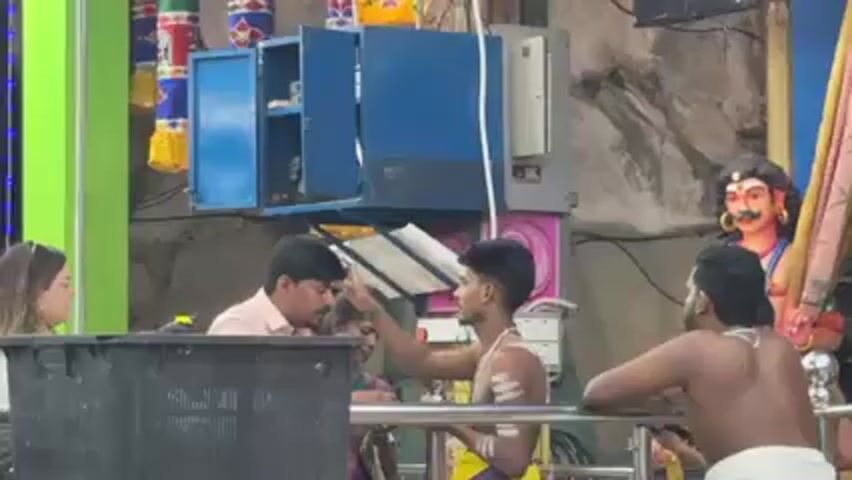 The video shows people inside a Hindu temple. There are men and women present, with some men wearing traditional clothing and shaved torsos.  A blue cabinet is visible behind them.  Statues and decorative elements can be seen, along with electrical wiring.  The people are engaged in various activities, including prayer.  The environment appears to be a bustling, religious setting.