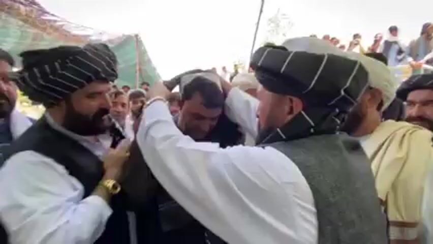 Here is a description of the scene:

The video shows a group of men gathered together, with three men in the foreground receiving the most attention. All three men are wearing traditional clothing. The man in the center is being adorned with a black and white scarf by one of the other men, who is wearing a gray vest over a white shirt. The man to the left is wearing a black vest over a white shirt. All three men have dark beards. The background is filled with other men, some wearing turbans and traditional clothing. There appears to be a tent structure visible in the background, suggesting an outdoor gathering. The environment seems to be a rural or less developed area, based on the tent and the clothing of the people present. The focus of the video seems to be on the man being dressed, indicating a possible ceremony or tradition being observed.