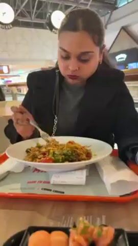 A woman is eating lunch in what appears to be a food court. She's dressed in a black coat and is using a fork to eat from a white plate filled with noodles, broccoli, tomatoes, and other ingredients. The plate has the logo of "Wok to Walk" on it. Next to her plate is a tray with a receipt. In the same tray is a black plate filled with sushi rolls and salmon nigiri. The sushi rolls are a mix of avocado, mango, and salmon. The food court is bright and bustling, with other people visible in the background.