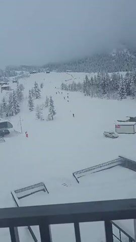 The video shows a snowy ski slope on a cloudy day. There are several skiers descending the hill. The landscape includes snow-covered trees and small buildings scattered throughout the area. In the foreground, there is a metal railing, indicating the viewpoint is from an elevated position. Towards the middle-left of the frame, a ski lift mechanism is visible. The overall scene depicts a winter recreational environment.