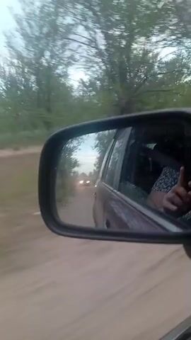 The video shows a view from inside a car driving on a dirt road. A side mirror reflects the road behind, showing another car with its headlights on and trees lining the road. A person's hand is visible in the reflection, giving a thumbs-up. The camera pans slightly, showing more of the dusty road and the surrounding vegetation. The sky appears overcast.