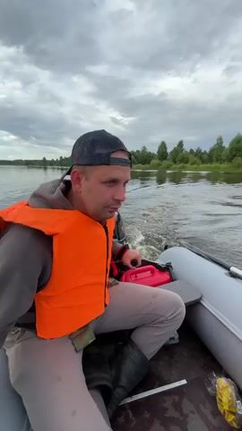The video shows a man wearing an orange life vest and a baseball cap. He is sitting in an inflatable boat on a body of water. The man is looking at the camera and making a hand gesture. There is a red gas can behind him. The boat is moving, creating a wake. The environment appears to be a river or lake with trees and a cloudy sky in the background. The man appears to be speaking or reacting to something off-camera.