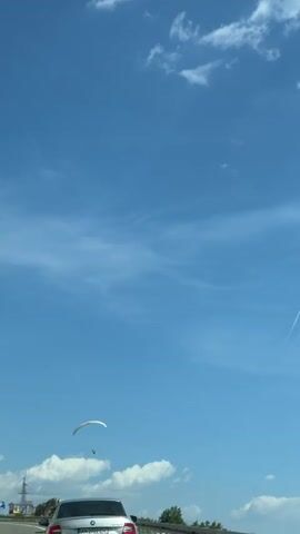 A paraglider is visible in the sky above a highway. A silver car is driving on the highway, and other vehicles are also present. The sky is blue with some clouds, and there are trees and a guardrail visible along the side of the road.