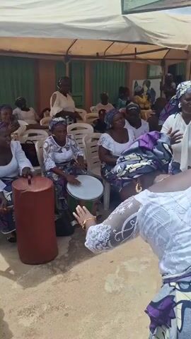 A group of women are dancing and playing instruments under a tent. The woman in the foreground is wearing a white lace top and a purple and gray patterned skirt with a matching headwrap. She is dancing with her back to the camera, bending forward and then standing upright. In the background, other women are also dancing and playing drums. One woman is playing a tall, reddish-brown drum, and another is playing a white-sided drum. The women are all dressed in similar white tops and patterned skirts.