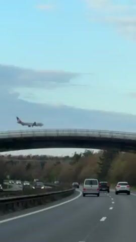 The video shows a view from inside a car driving on a highway. The highway has multiple lanes, with cars and a van in the lanes ahead. An overpass bridge is visible, and an airplane is seen flying low, passing behind the bridge. Trees line the side of the road, and the sky is partially cloudy.
