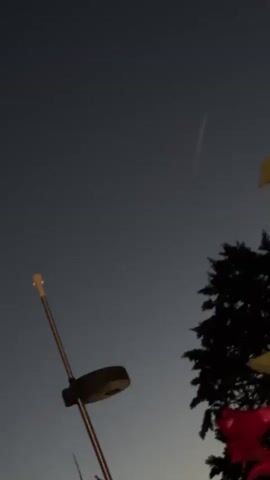 The video shows a dark sky with a few faint lights. In the foreground, a metal pole with a light fixture is visible on the left. On the right, the silhouette of a tree is seen with some colorful objects, possibly flags or decorations, in front of it. A faint streak of light, possibly a meteor or satellite, is visible in the upper right quadrant of the sky. The overall lighting suggests it is either dusk or pre-dawn.