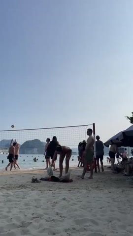 This is a vertical, medium-wide shot of a beach scene. In the foreground, there is sand with a few items of clothing scattered on it. A volleyball net is set up on the sand, with a volleyball in the air over the net. A group of people are playing volleyball, and several others are standing around the net. In the midground, there is the ocean with several people swimming and standing in the water. In the background, there are several large, lush green islands with boats in the water around them. The sky is clear and blue. The overall mood is relaxed and recreational.