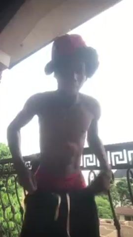 A shirtless young man with curly hair and a red cap dances on a balcony with an ornate metal railing. The sky is overcast, and trees are visible in the background. The man gestures and laughs as he dances.