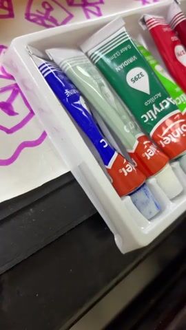 A close-up shot shows a set of acrylic paints in a white plastic tray with individual compartments for each tube. The tubes are mostly standing upright, with the bottom end visible.  The paint colors visible are shades of blue, green, red, orange and yellow.  A few of the paint tube caps have dabs of their respective colors.  The brand name "Finilico Acrilico Pointer" is seen on the tubes.  Around the tray are various other art-related items including a yellow paint palette with green paint, a box of watercolor pencils and a towel. A sheet of paper with a drawing in purple ink is also present.  The surface underneath appears to be a dark-colored table. The camera moves slightly, providing different angles of the paint set.